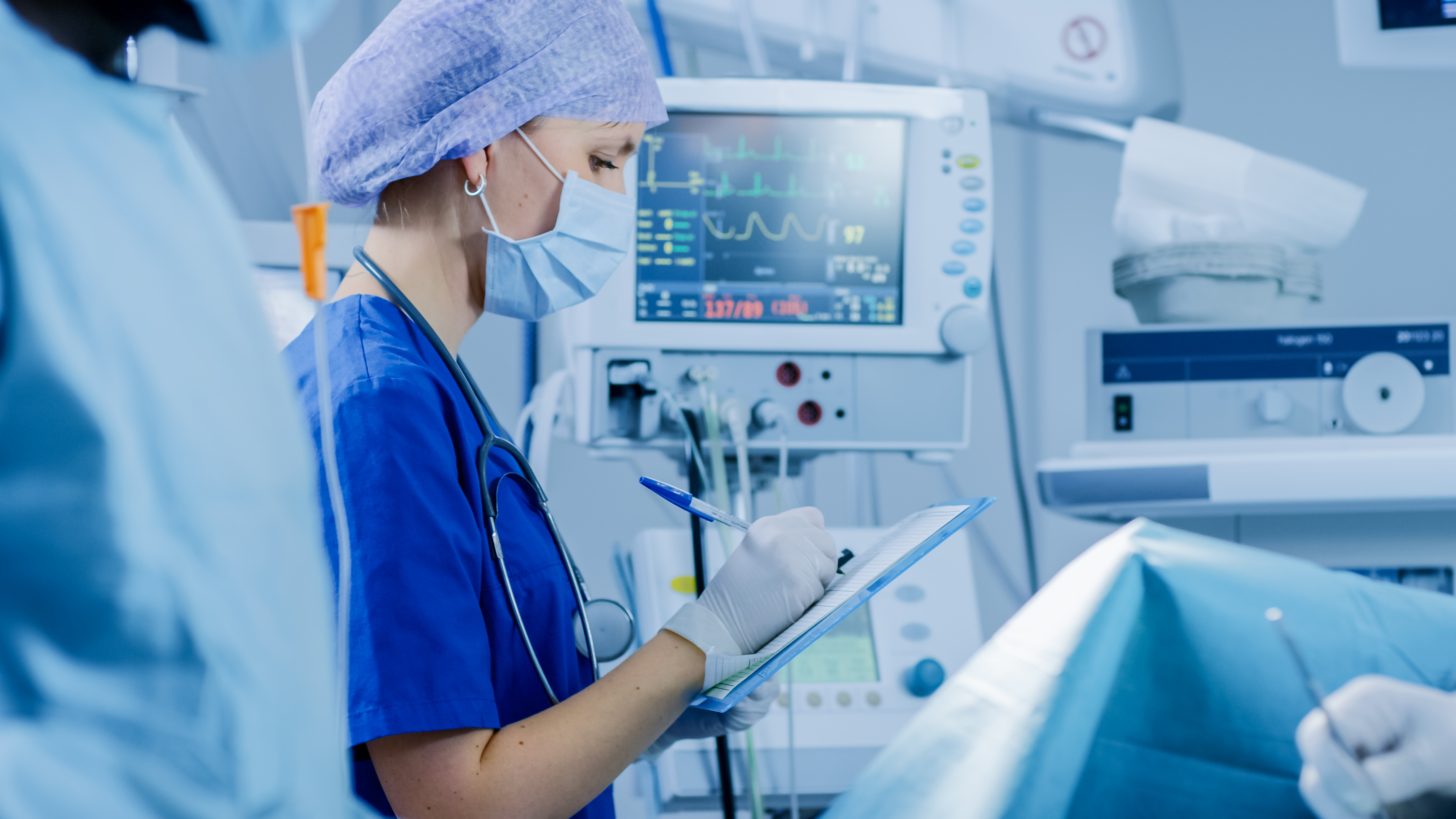 Medical staff taking notes on a chart in a hospital room with medical devices in the background.